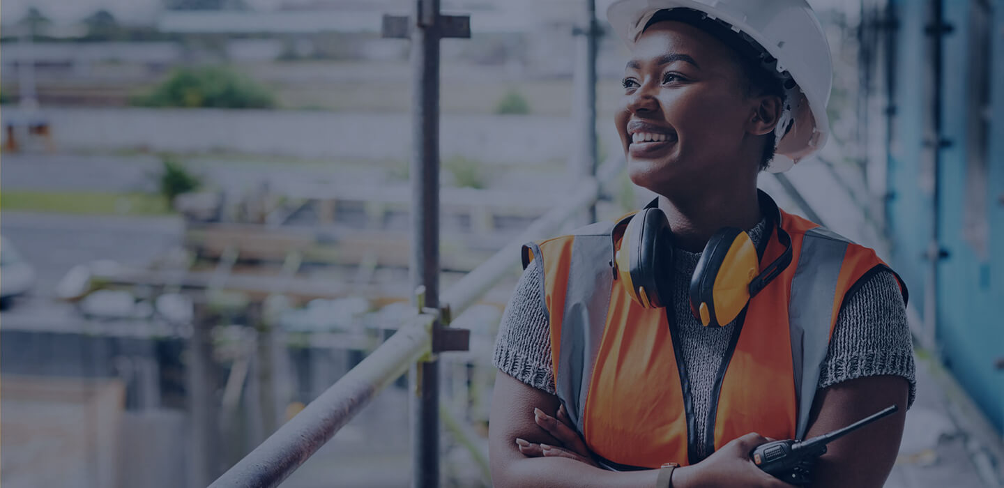 Smiling woman on construction site, wearing orange vest and hard hat, holding walkie talkie