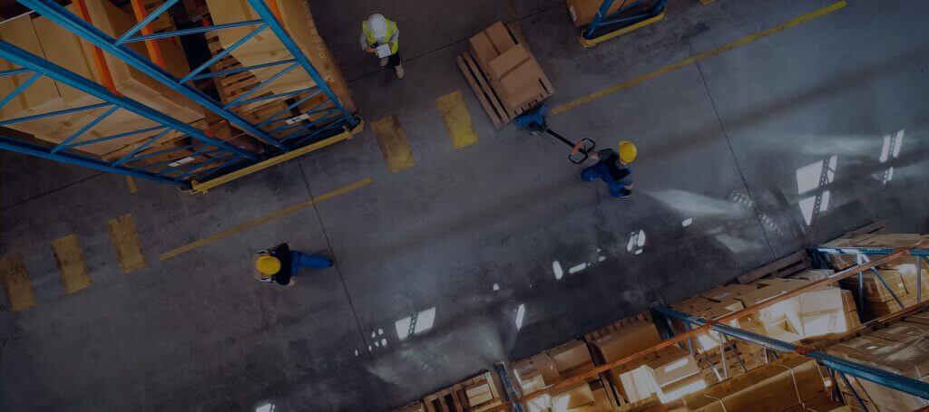 A photo of warehouse workers taken from above, one worker is moving a hand-powered forklift with boxes and another is walking nearby